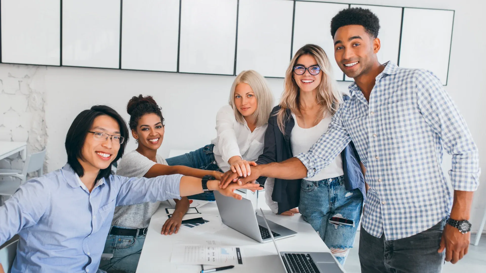  A diverse group of five business professionals in casual clothing smiling and holding their hands together over a desk in a modern office space.